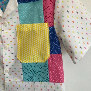 May include: A white short-sleeved button-up shirt with a patchwork design. The shirt features different colored fabrics with polka dot patterns in yellow, blue, pink, and green. The shirt has a yellow pocket with white polka dots.