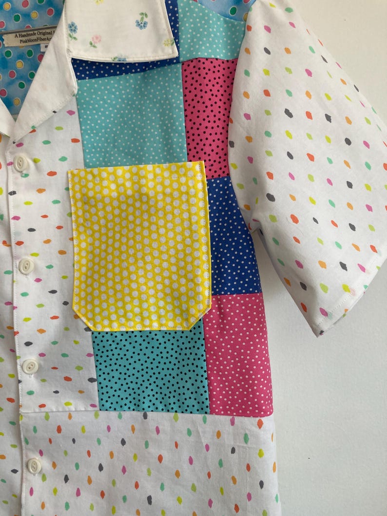 May include: A white short-sleeved button-up shirt with a patchwork design. The shirt features different colored fabrics with polka dot patterns in yellow, blue, pink, and green. The shirt has a yellow pocket with white polka dots.