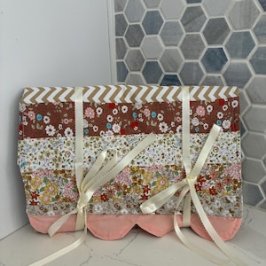 May include: A rectangular fabric organizer featuring horizontal stripes of various floral patterns in brown, white, pink, and yellow. The top edge has a chevron print, and the bottom edge is scalloped in solid pink. Two cream ribbons tie the front.