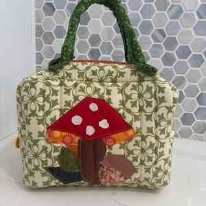 May include: A quilted fabric handbag featuring a cream and green damask pattern with a large red mushroom appliqué on the front. The bag has a green patterned handle and a zipper closure. The mushroom design includes white spots and brown stems.