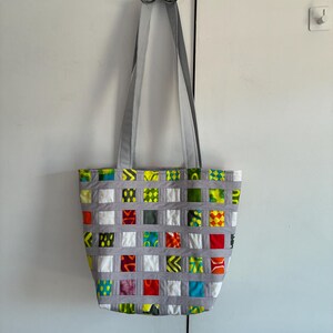 May include: A tote bag featuring a grid pattern created by grey fabric strips layered over a patchwork of colorful fabric squares. The squares display various patterns and colors including red, yellow, green, blue, and white. It has two grey straps.