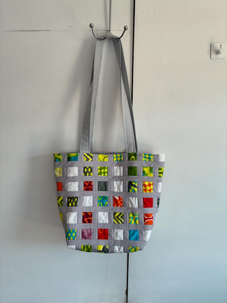 Op de afbeelding: Een tote bag met een ruitpatroon gecre&euml;erd door grijze stoffen stroken over een patchwork van kleurrijke stoffen vierkantjes. De vierkantjes tonen diverse patronen en kleuren, waaronder rood, geel, groen, blauw en wit. Het heeft twee grijze hengsels.