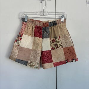 May include: Patchwork shorts in a variety of colors, including beige, red, and gray, hang from a clear plastic hanger. The shorts are made from squares of fabric sewn together, creating a quilt-like pattern. The elastic waistband is visible.