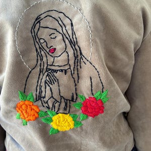 May include: A tan jacket featuring an embroidered design of a woman with a halo, hands clasped in prayer, and a floral garland. The embroidery includes black, red, orange, yellow, and green thread. The jacket is made of a soft, suede-like material.