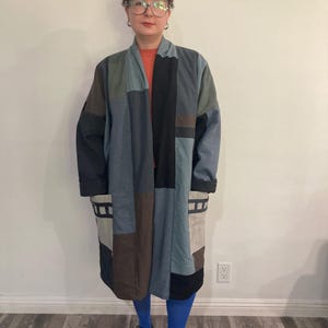 May include: A long, patchwork coat in shades of blue, black, and brown. The coat has a relaxed fit and a collarless design. The person wearing the coat is also wearing blue tights and black shoes.