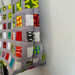 Op de afbeelding: Een draagtas met een geweven rasterontwerp, bestaande uit grijze stoffen stroken die zijn verweven met vierkanten van veelkleurige bedrukte stof. De vierkanten tonen diverse geometrische en abstracte prints in tinten rood, geel, groen, blauw en wit.