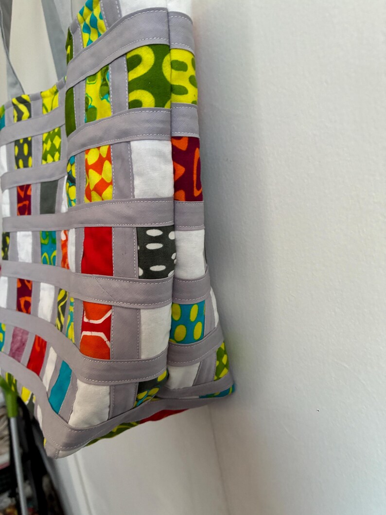 Op de afbeelding: Een draagtas met een geweven rasterontwerp, bestaande uit grijze stoffen stroken die zijn verweven met vierkanten van veelkleurige bedrukte stof. De vierkanten tonen diverse geometrische en abstracte prints in tinten rood, geel, groen, blauw en wit.