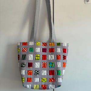 Op de afbeelding: Een tote bag met een geweven rasterontwerp van lichtgrijze stoffen stroken. Het raster bevat kleine, vierkante stoffen lapjes in diverse felle kleuren en patronen, waaronder rood, oranje, geel, groen, wit en zwart, voorzien van twee lange hengsels.