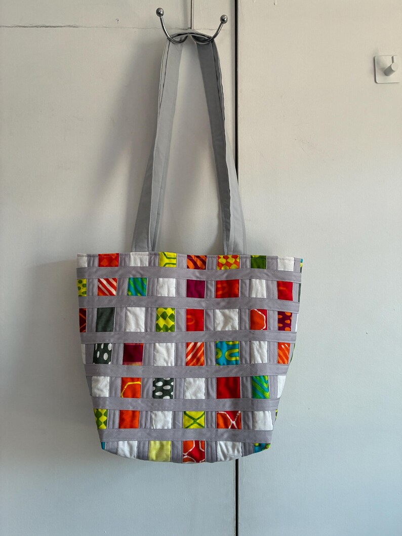 Op de afbeelding: Een tote bag met een geweven rasterontwerp van lichtgrijze stoffen stroken. Het raster bevat kleine, vierkante stoffen lapjes in diverse felle kleuren en patronen, waaronder rood, oranje, geel, groen, wit en zwart, voorzien van twee lange hengsels.