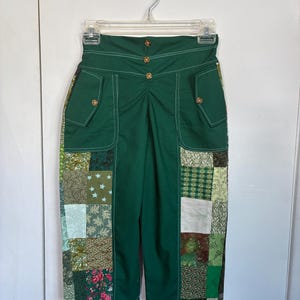 May include: Green high-waisted pants with a patchwork design on the sides. The pants have a button closure and pockets. The patchwork features various patterns in shades of green, brown, and white. The pants are hanging on a clear hanger.