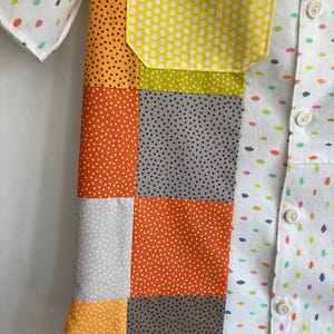 May include: A white short-sleeved shirt with a patchwork design on the front. The patchwork is made up of different colored fabrics with white polka dots. The shirt has a button-up closure.