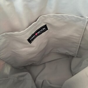 Op de afbeelding: De binnenkant van een tote bag met een lichtgrijze stoffen voering en een open vak. Een zwart rechthoekig stoffen label is op het vak genaaid, met de witte tekst PINK MOON en een kleine roze cirkel tussen de twee woorden.
