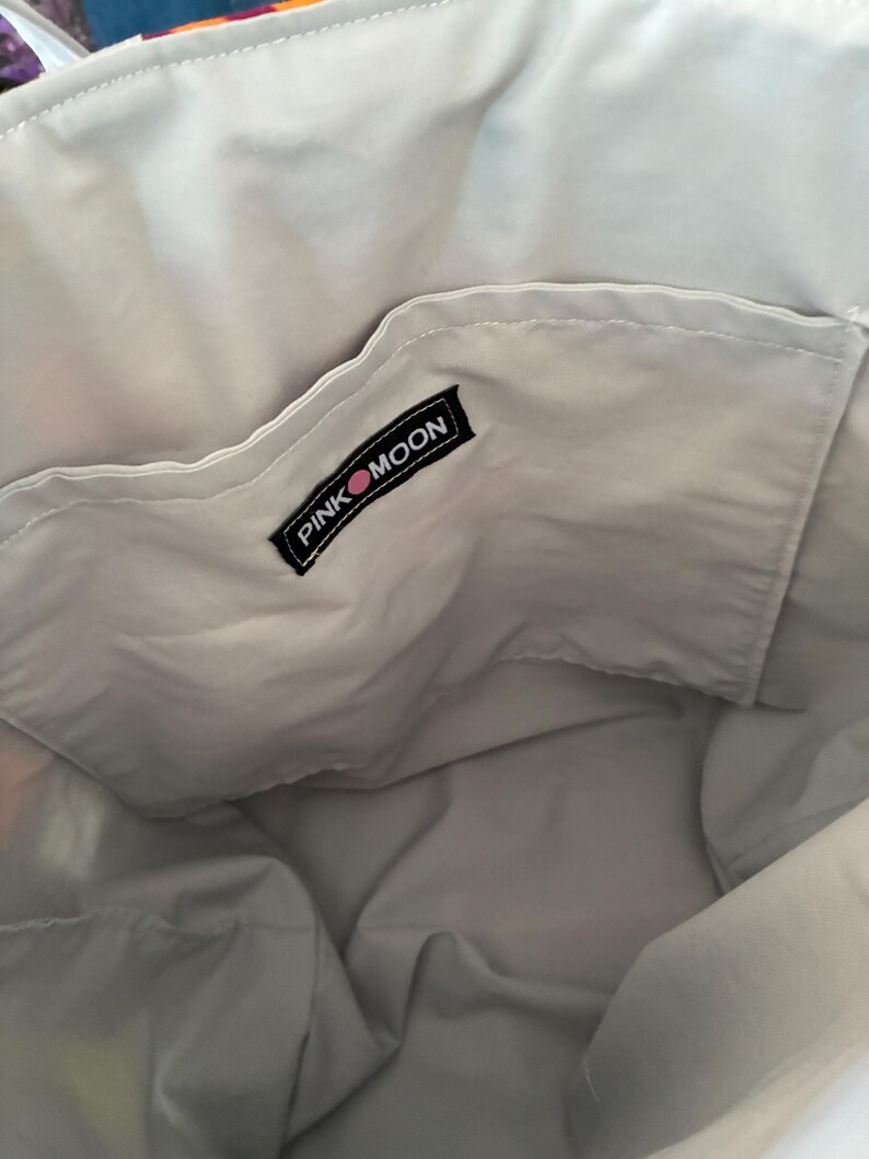 Op de afbeelding: De binnenkant van een tote bag met een lichtgrijze stoffen voering en een open vak. Een zwart rechthoekig stoffen label is op het vak genaaid, met de witte tekst PINK MOON en een kleine roze cirkel tussen de twee woorden.