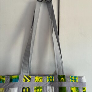 Op de afbeelding: Een tote bag met een patchworkontwerp van diverse groene, gele, witte en oranje stoffen vierkanten. De tas is geconstrueerd met lichtgrijze stoffen stroken die een rasterpatroon vormen over de vierkanten en heeft twee lange grijze schouderbanden.