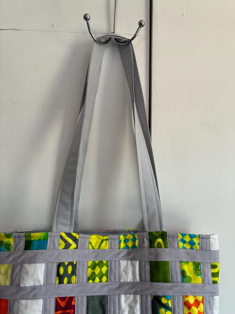 Op de afbeelding: Een tote bag met een patchworkontwerp van diverse groene, gele, witte en oranje stoffen vierkanten. De tas is geconstrueerd met lichtgrijze stoffen stroken die een rasterpatroon vormen over de vierkanten en heeft twee lange grijze schouderbanden.