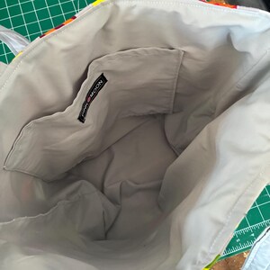 Op de afbeelding: Een tote bag met een lichtgrijze voering en een open binnenzak. Een zwart stoffen label met de witte tekst PINK MOON is op de bovenrand van de zak genaaid. De buitenrand is voorzien van een kleurrijke stof met een patroon.