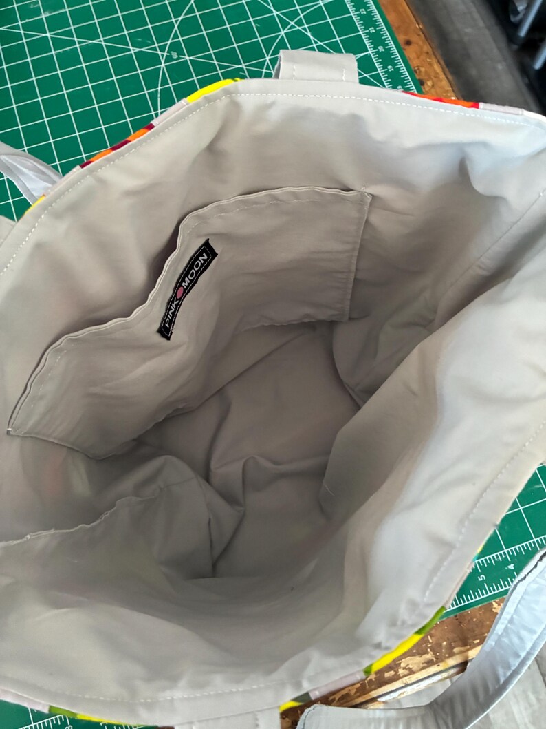 Op de afbeelding: Een tote bag met een lichtgrijze voering en een open binnenzak. Een zwart stoffen label met de witte tekst PINK MOON is op de bovenrand van de zak genaaid. De buitenrand is voorzien van een kleurrijke stof met een patroon.