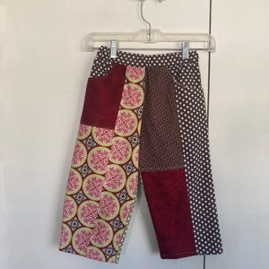 May include: Patchwork pants with a variety of patterns and colors. The pants feature a mix of fabrics, including a burgundy solid, a floral print with red and green accents, a brown floral, and a white polka dot pattern. The pants are hanging on a clear hanger.