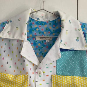 May include: A white button-up shirt with a patchwork design featuring different colored polka dots and floral patterns. The shirt has a collar and a pocket on the left side. The label reads "PinkMoonFiberArts Original by".