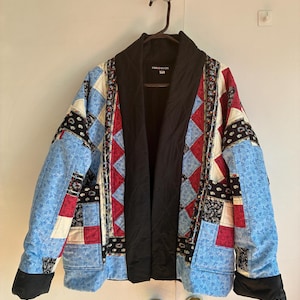 May include: A quilted jacket featuring a patchwork design of blue floral, red, and black paisley fabric squares. The garment has a solid black shawl collar, black sleeve cuffs, and a black interior lining. The brand label reads PINK MOON.