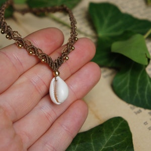 Könnte beinhalten: Eine braune und beigefarbene Makramee-Kette mit goldenen Perlen und einem weißen Kaurischnecken-Anhänger.