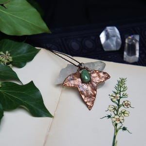 May include: A copper leaf pendant necklace with a green gemstone in the center. The necklace is displayed on a book with botanical illustrations.