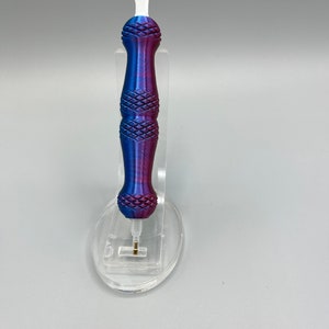 May include: A purple and blue pen with a white tip and a diamond pattern on the handle. The pen is sitting on a clear plastic stand.