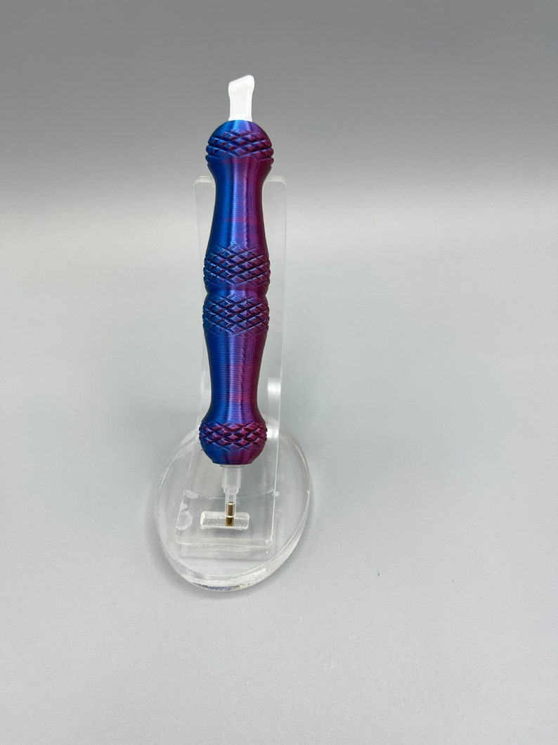 May include: A purple and blue pen with a white tip and a diamond pattern on the handle. The pen is sitting on a clear plastic stand.
