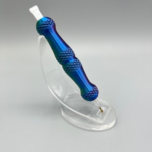 May include: A blue and purple metal tattoo pen with a white tip, sitting on a clear plastic stand.