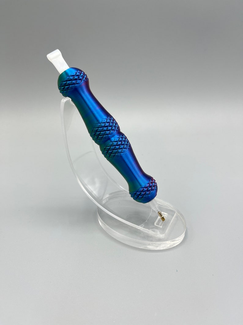 May include: A blue and purple metal tattoo pen with a white tip, sitting on a clear plastic stand.