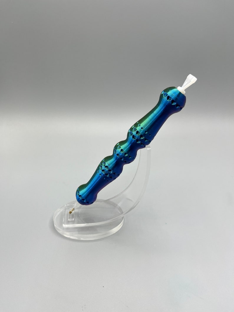 May include: A blue and green iridescent pen with a textured handle and a white tip. The pen is on a clear plastic stand.
