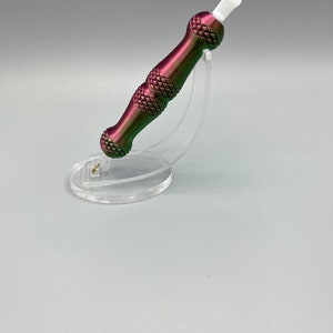 May include: A purple and green metal tool with a white tip, sitting on a clear plastic stand. The tool has a textured, diamond-shaped pattern.