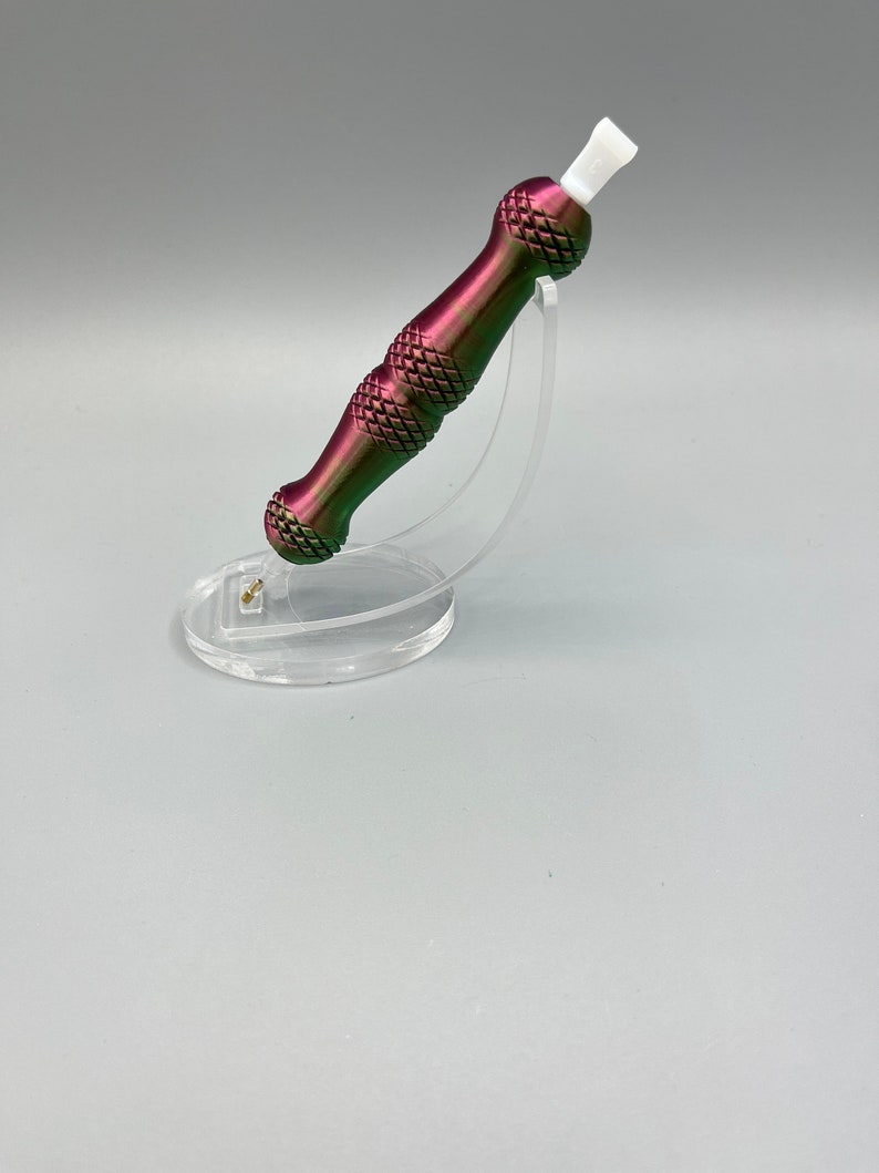 May include: A purple and green metal tool with a white tip, sitting on a clear plastic stand. The tool has a textured, diamond-shaped pattern.