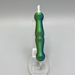 May include: A green and blue iridescent pen with a white tip and a diamond pattern on the barrel. The pen is on a clear plastic stand.