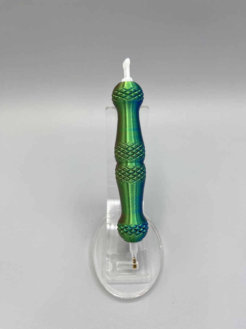 May include: A green and blue iridescent pen with a white tip and a diamond pattern on the barrel. The pen is on a clear plastic stand.