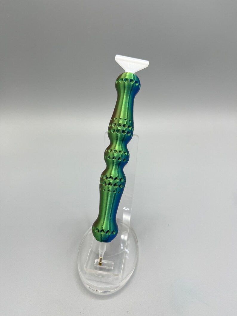 May include: A green, blue, and white diamond painting tool with a textured handle. The tool is made of plastic and has a small, pointed tip.