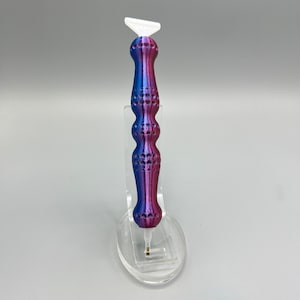 May include: A purple and blue diamond painting pen with a white tip. The pen has a textured handle and is displayed on a clear stand.