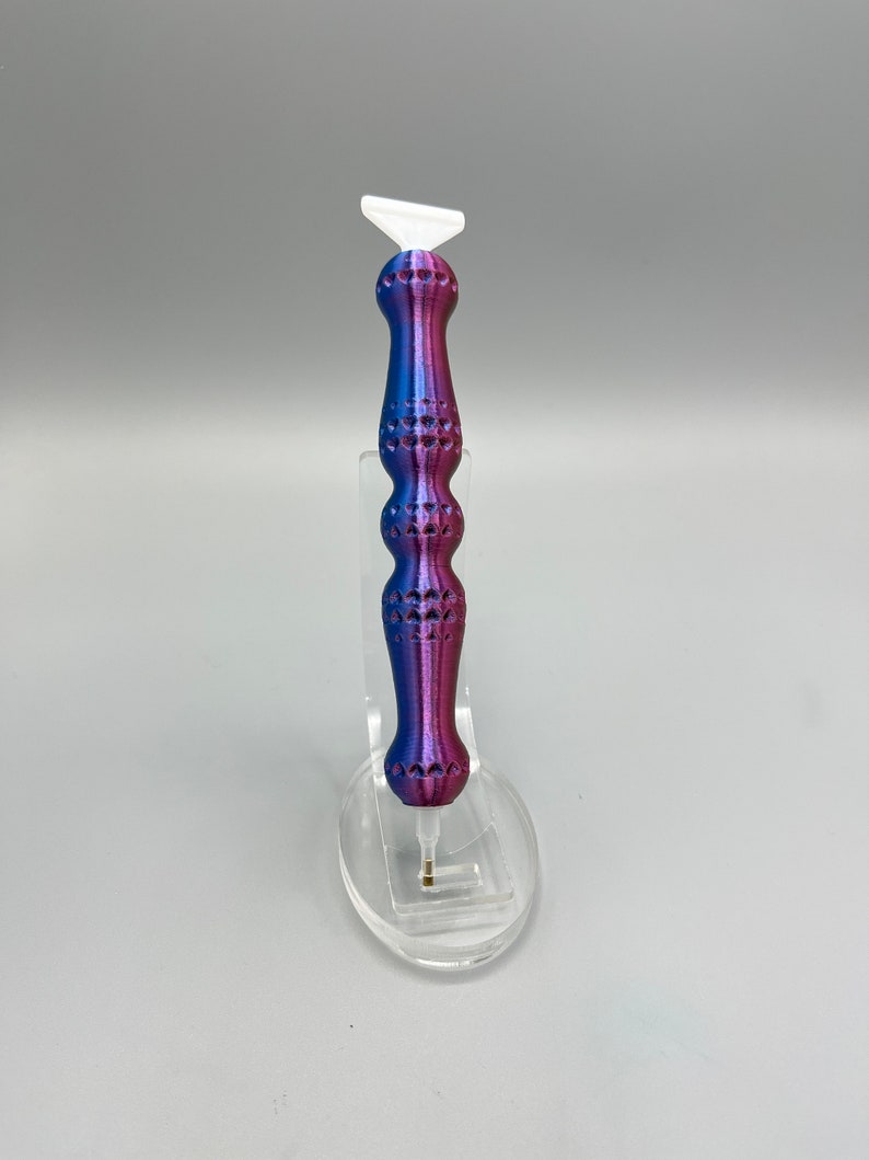 May include: A purple and blue diamond painting pen with a white tip. The pen has a textured handle and is displayed on a clear stand.