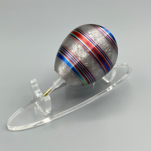 May include: A silver egg with colorful stripes on a clear acrylic stand. The egg has a textured surface and is decorated with blue, red, and green stripes.