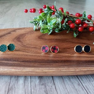 May include: Three pairs of stud earrings on a wooden surface. The earrings are made of gold metal and have different patterns: green, red and black plaid, and black.