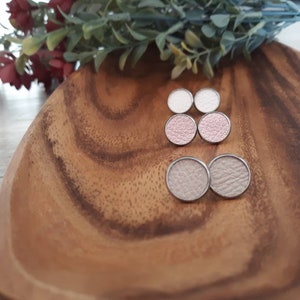 May include: Three pairs of round leather earrings in white, light pink, and dark pink. The earrings are displayed on a wooden surface.