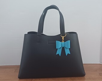 May include: Black tote bag with two handles and a light blue bow-shaped charm. The bag has a rectangular shape and a simple design. The bow charm is attached to the handle with a gold-colored clasp.