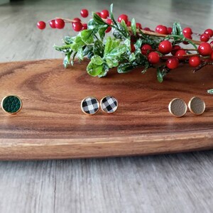 May include: Three pairs of small, round stud earrings on a wooden surface. The earrings are gold-toned and feature different patterns: green, black and white checkered, and brown.
