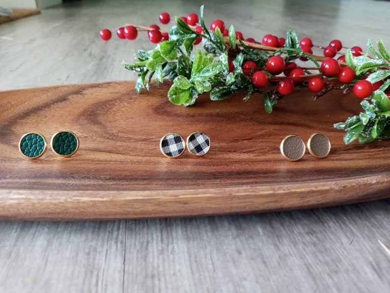 May include: Three pairs of small, round stud earrings on a wooden surface. The earrings are gold-toned and feature different patterns: green, black and white checkered, and brown.