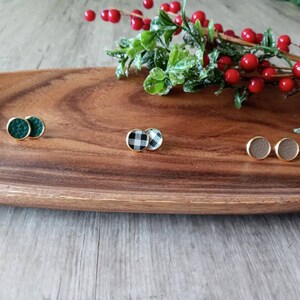 May include: Three pairs of small, round, gold-toned earrings with different patterns. The earrings are on a wooden surface. The patterns are green, black and white checkered, and brown.