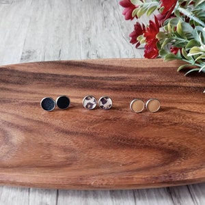 May include: Three pairs of small round earrings on a wooden tray. The earrings are made of metal and have different fabric patterns: black, leopard print, and tan.