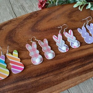 May include: A set of six pairs of Easter bunny-shaped earrings. The earrings are made of faux leather and feature various patterns, including stripes, polka dots, and gingham. Each pair has a small white pom-pom on the back.
