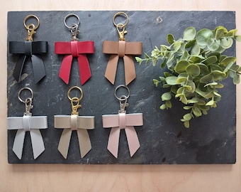 May include: Six leather keychains with bow designs in various colors, including black, red, brown, silver, beige, and pink, displayed on a dark gray surface.