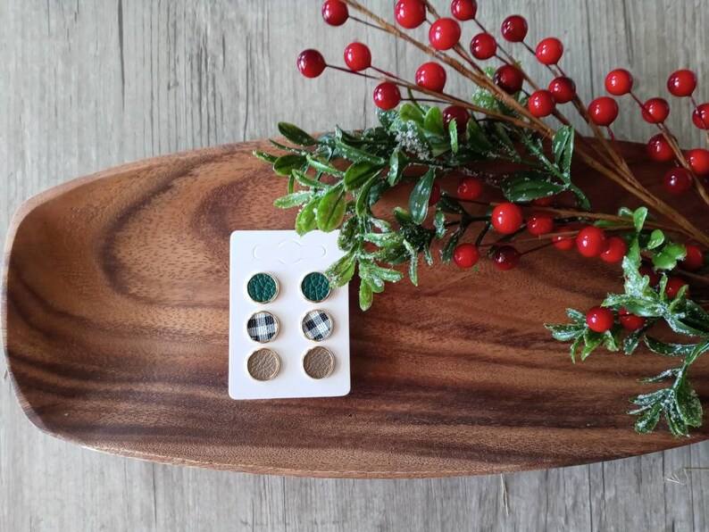 May include: A set of six small round earrings on a white card. The earrings are made of leather and have a green, black and white checkered, and brown design.