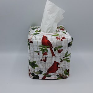 May include: A square tissue box cover featuring a festive design. The cover is adorned with red cardinals perched on branches with red berries and green leaves, set against a backdrop of vintage sheet music. White tissues are visible at the top.